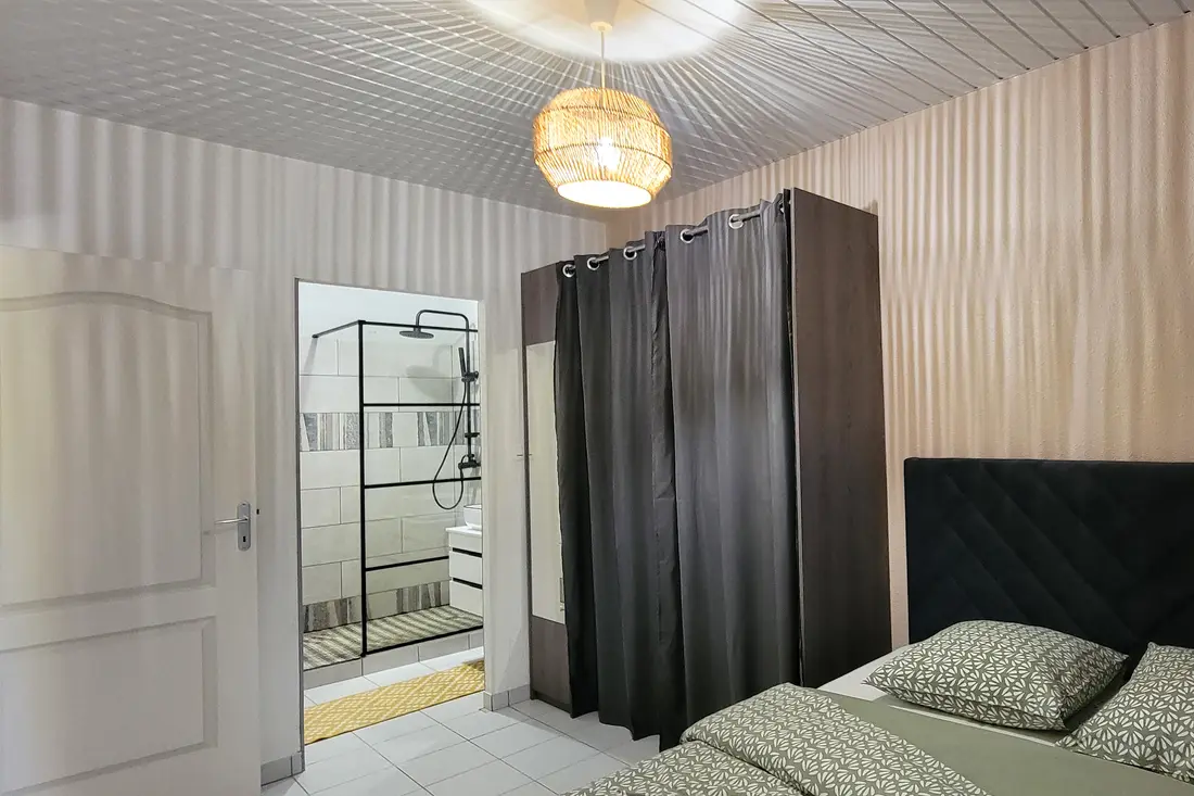 Chambre d’un appartement Cayenne Chic : tête de lit capitonnée, penderie à rideaux et salle d’eau attenante avec douche