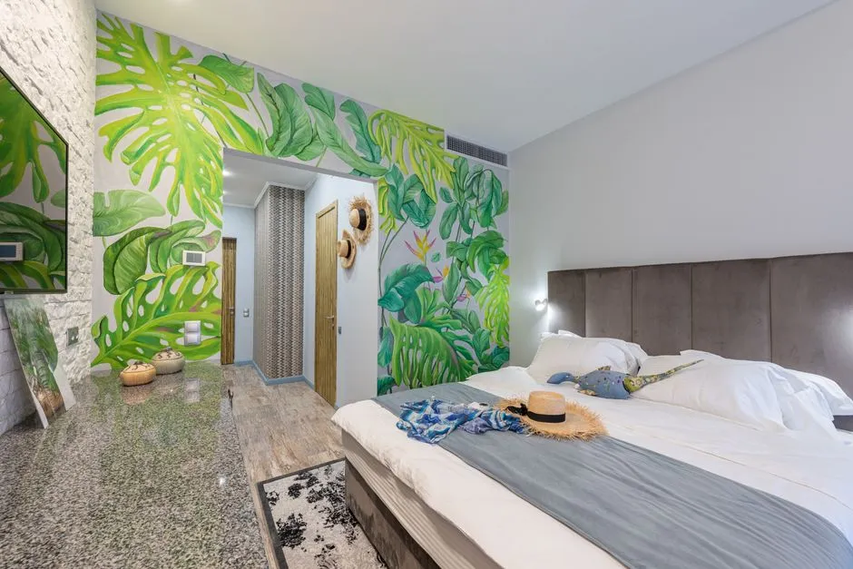 Chambre d'hôtel tropicale lumineuse avec mur végétal en feuilles de monstera, lit double et chapeau de paille