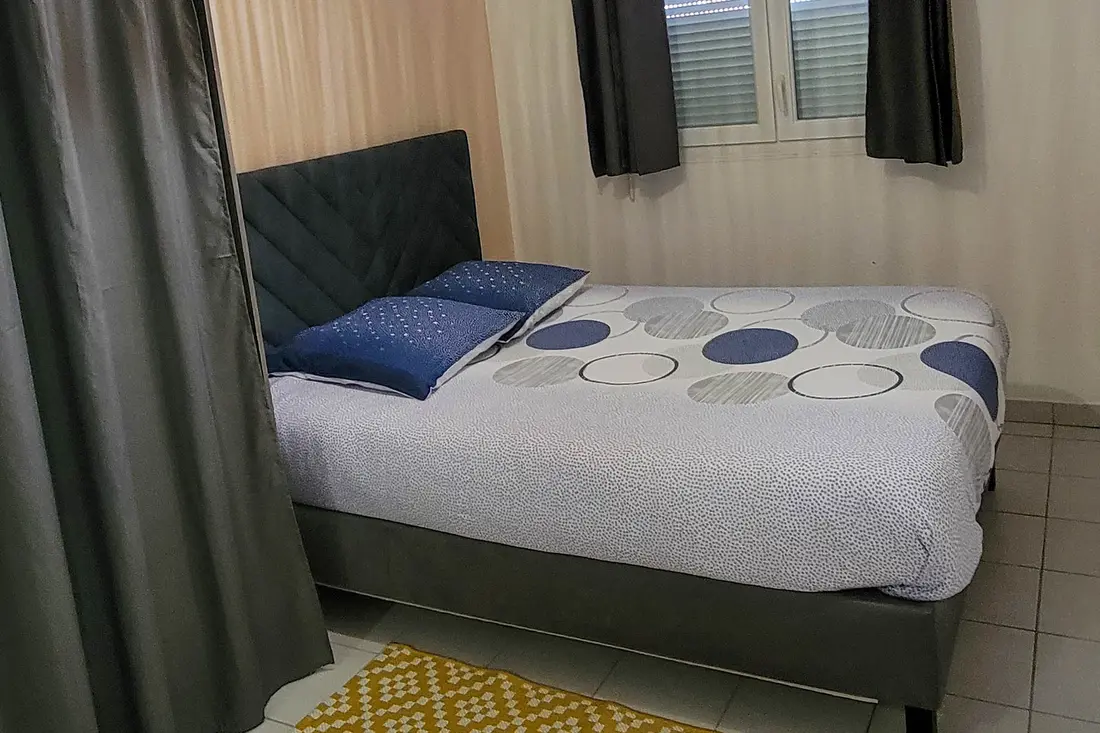 Chambre d’un appartement Cayenne Chic : lit à tête capitonnée grise, parure à motifs bleus, suspension en rotin et tapis jaune