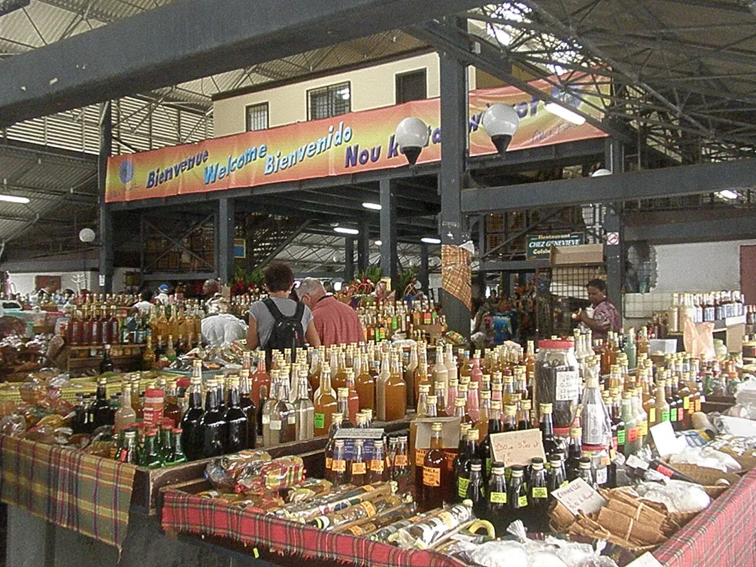 Étal de rhums arrangés et de produits locaux sous la halle du Grand Marché de Fort-de-France, avec une banderole de bienvenue en français, anglais, espagnol et créole