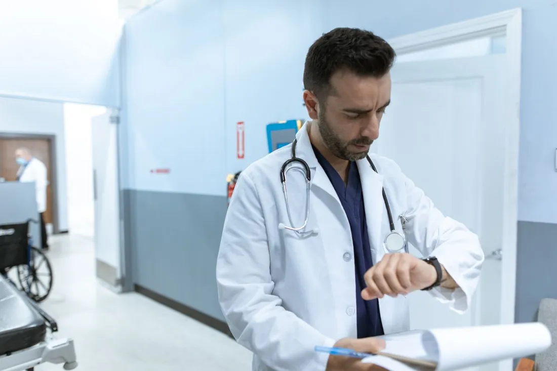 Un médecin en blouse blanche, stéthoscope autour du cou et dossier à la main, regarde sa montre dans un couloir d'hôpital