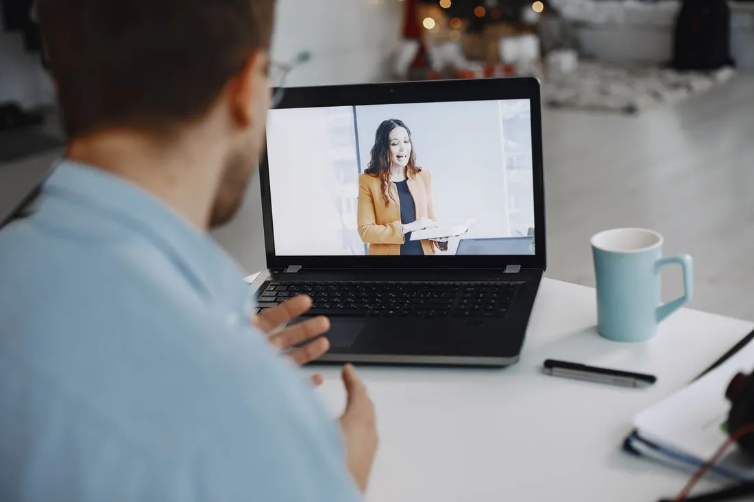Un homme assis à un bureau blanc suit une visioconférence sur un ordinateur portable, une tasse et un carnet posés à côté de lui