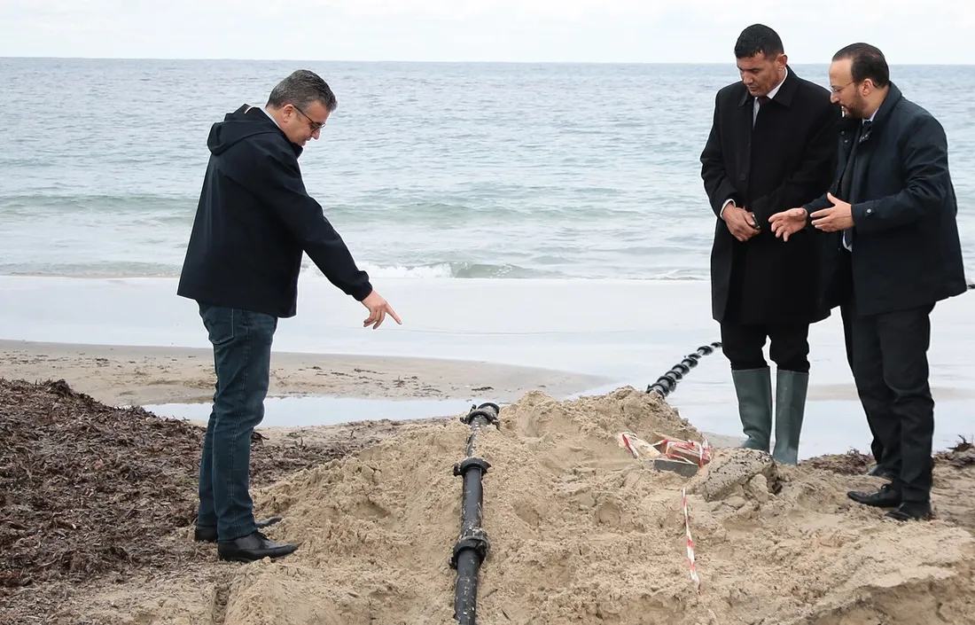 Trois hommes debout sur une plage observent un câble sous-marin noir sorti du sable et posé dans une tranchée jusqu'au bord de l'eau