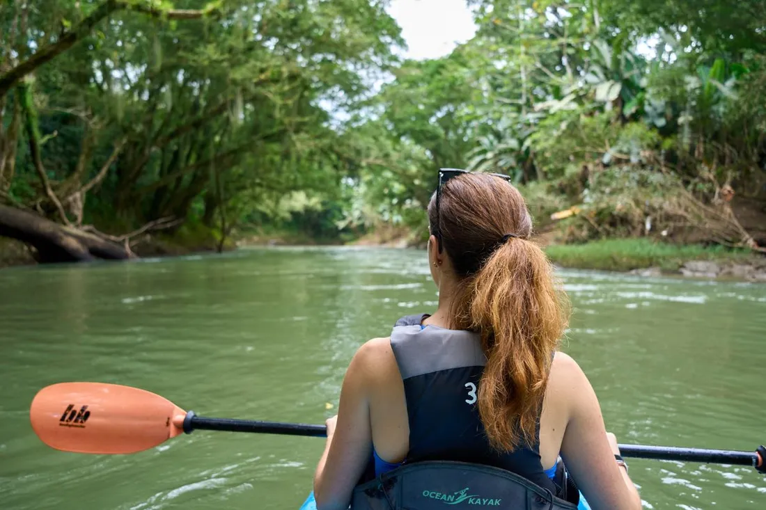 Kayakiste pagayant sur une crique bordee de foret tropicale dense, vue depuis l'arriere du kayak