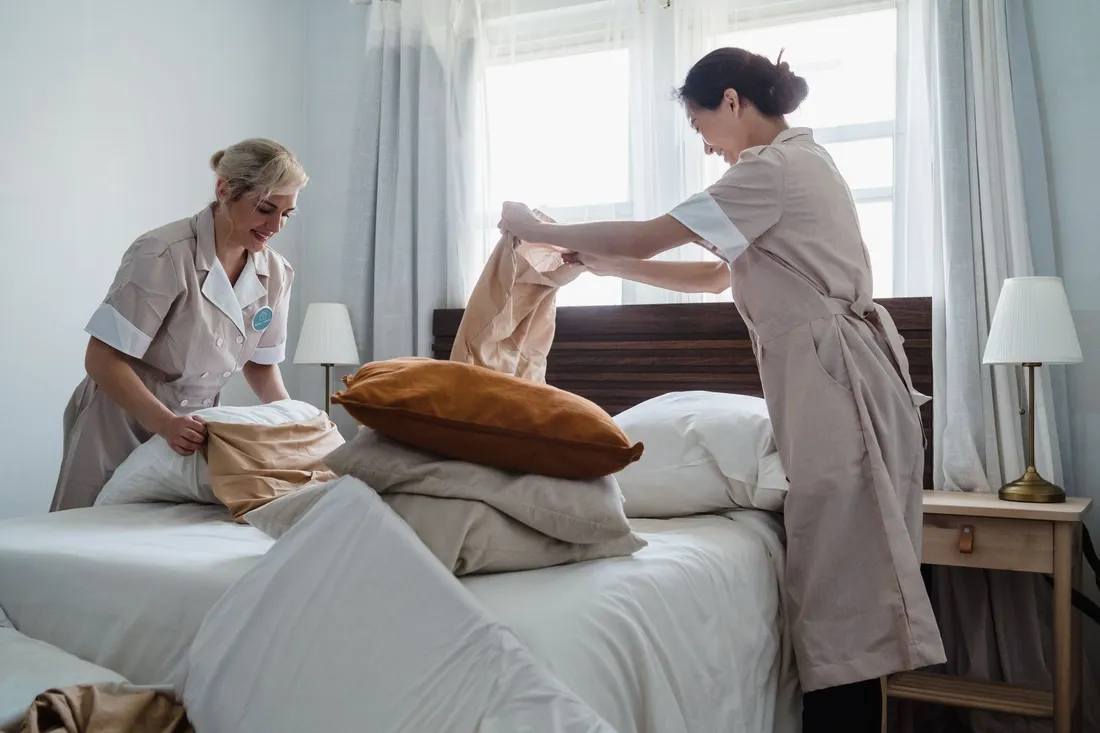 Deux employées de ménage en tenue beige refont un lit dans une chambre claire : l'une déplie une taie d'oreiller, l'autre en enfile une, draps blancs et coussins empilés