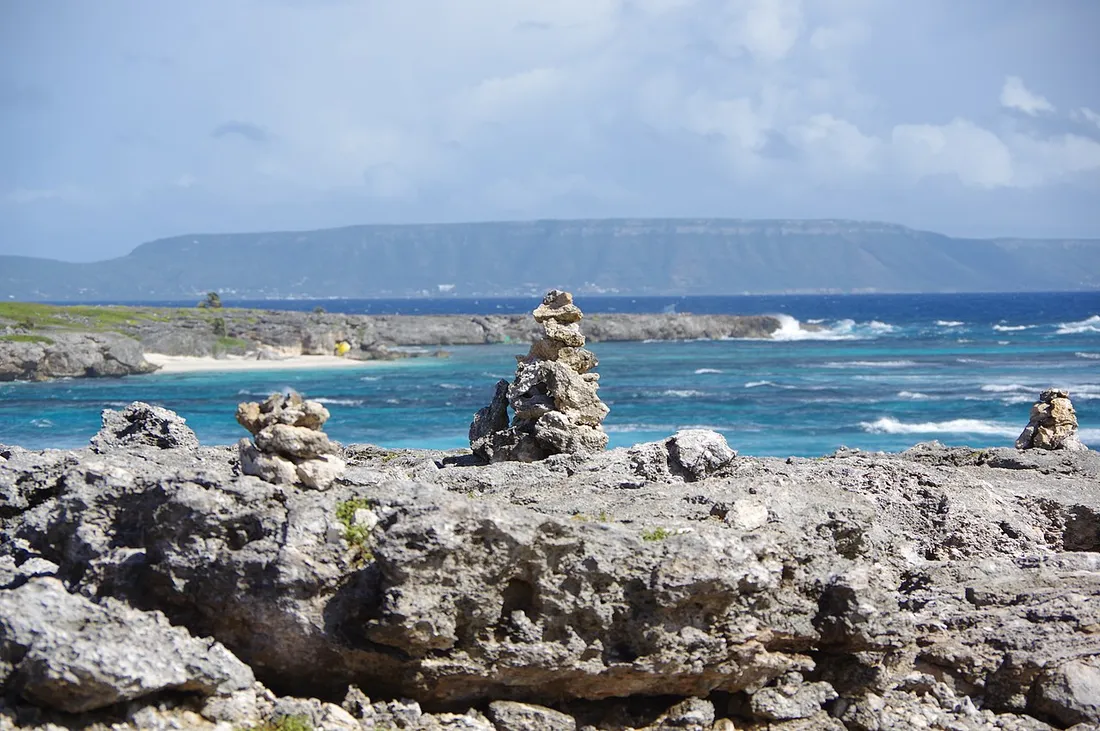 Le plateau calcaire de La Désirade vu depuis Petite-Terre, au-dessus d'une mer turquoise battue par les vagues, avec des cairns de pierres au premier plan