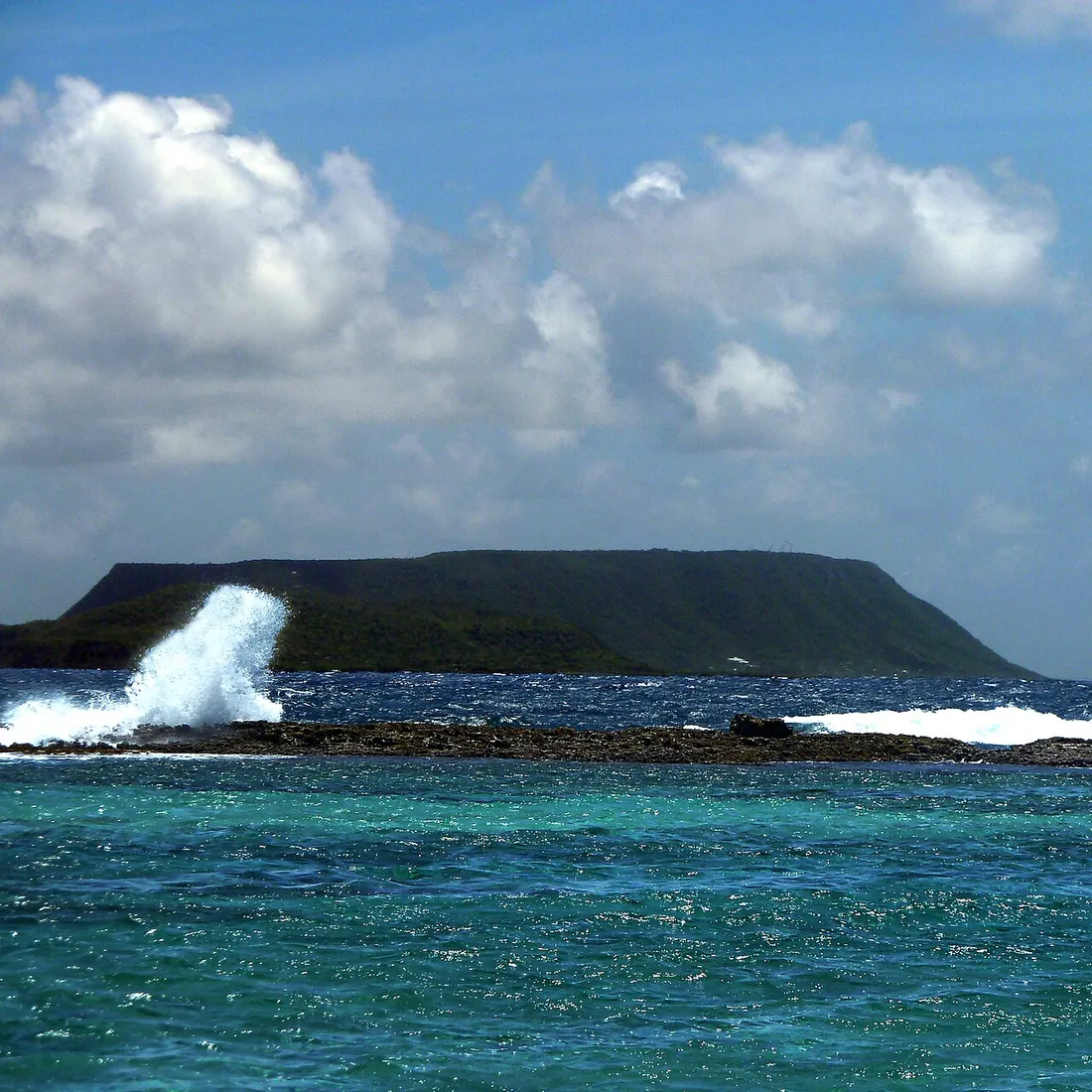 L'île de La Désirade et son plateau allongé vus depuis la mer des Caraïbes, avec une vague se brisant sur la barre rocheuse au large