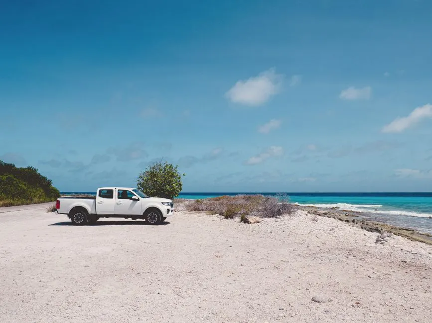 Pick-up de location blanc stationné sur une piste de sable au bord d'une mer turquoise des Caraïbes