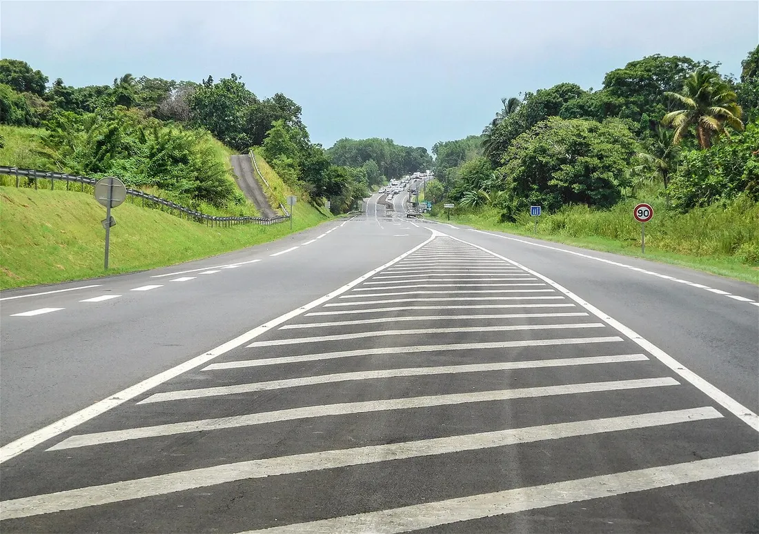 Route nationale de Guadeloupe bordee de vegetation tropicale avec panneau de limitation a 90 km/h, typique des trajets en voiture sur l'ile