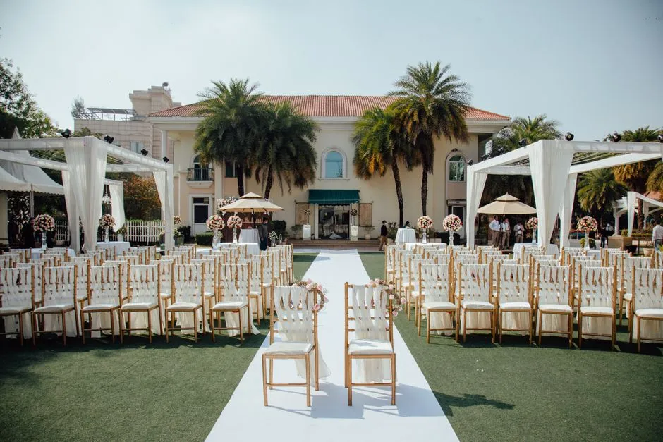 Cérémonie de mariage en extérieur dressée sur une pelouse devant une villa, avec allée blanche, chaises dorées et pergolas drapées sous les palmiers