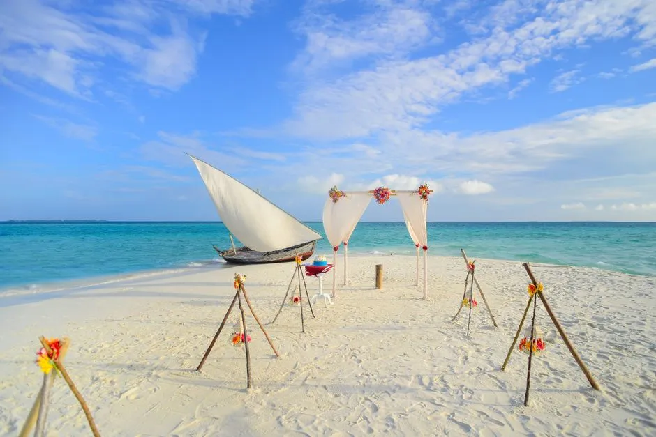 Decor de mariage installe sur une plage de sable blanc avec arche fleurie face a la mer turquoise et un voilier