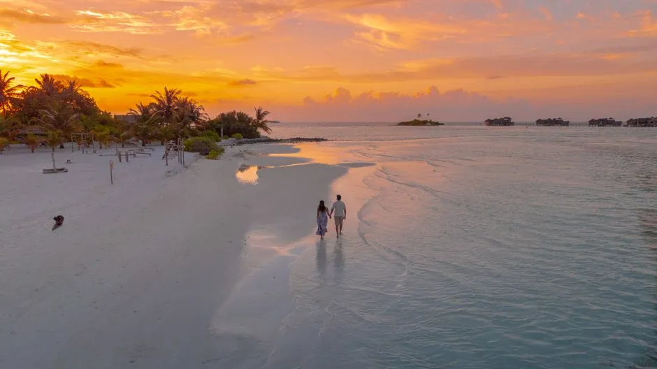 Couple de jeunes maries marchant main dans la main au bord de l'eau sur une plage tropicale au coucher du soleil