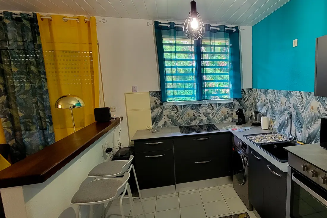 Cuisine d’un appartement Cayenne Chic : murs bleu vif et rideaux jaunes, meubles anthracite, bar en bois et tabourets