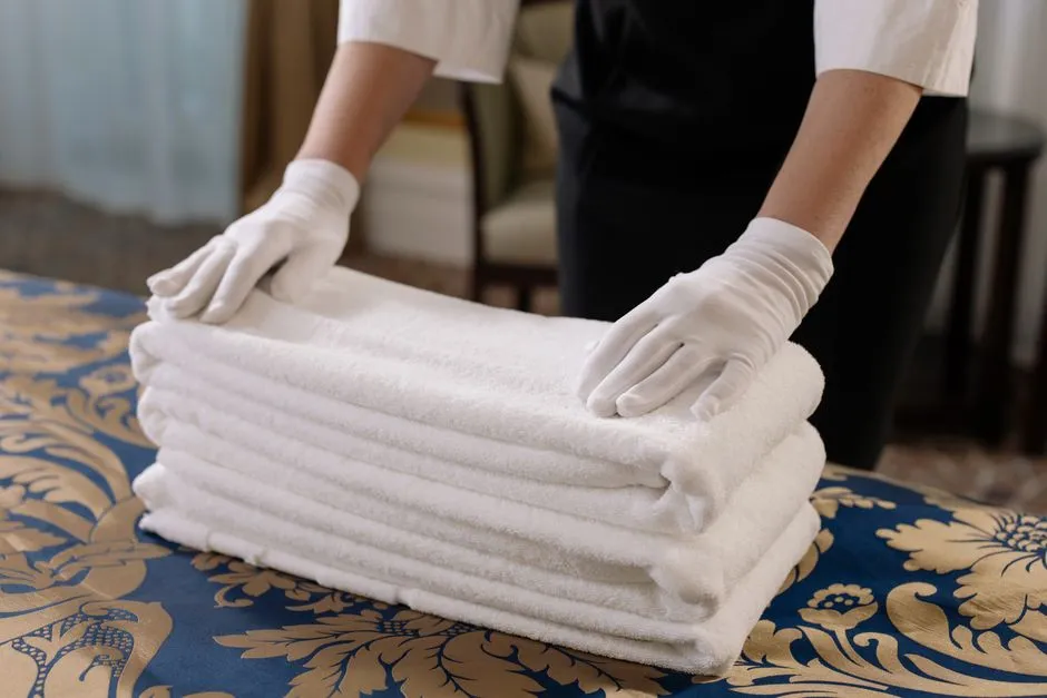 Femme de ménage portant des gants blancs pliant des serviettes éponge blanches propres sur un lit, gestion de la blanchisserie d'un logement de location