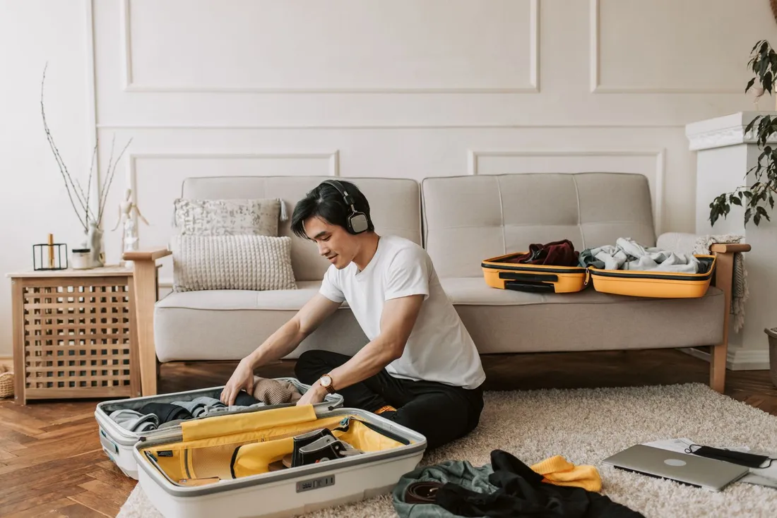 Un homme assis au sol dans un séjour plie des vêtements dans une grande valise ouverte, une seconde valise posée sur le canapé derrière lui