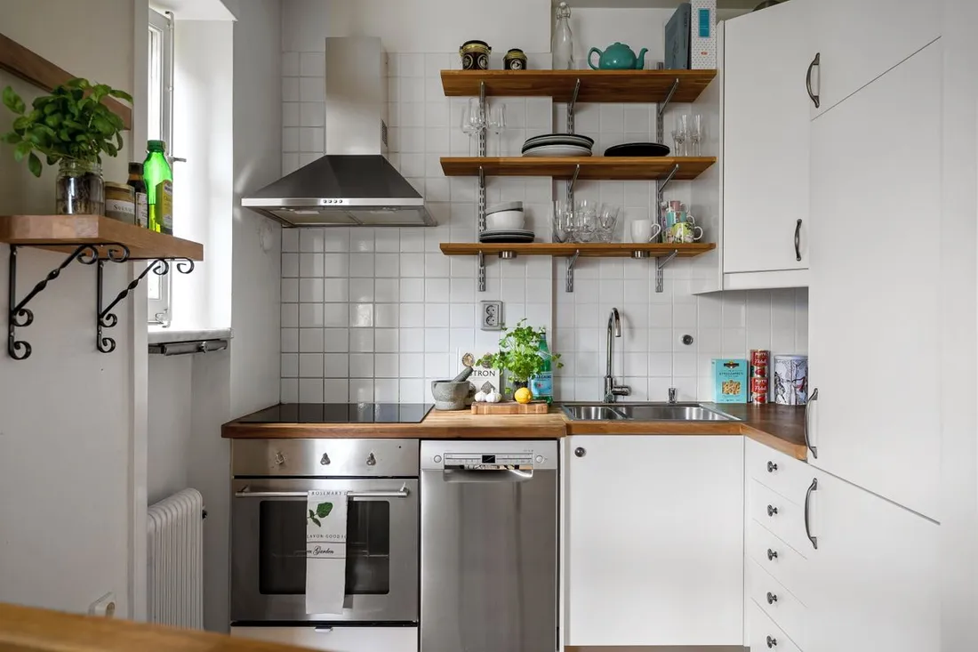Petite cuisine équipée d'un appartement : plaque de cuisson, four, lave-vaisselle, évier sous la fenêtre et étagères ouvertes garnies de vaisselle