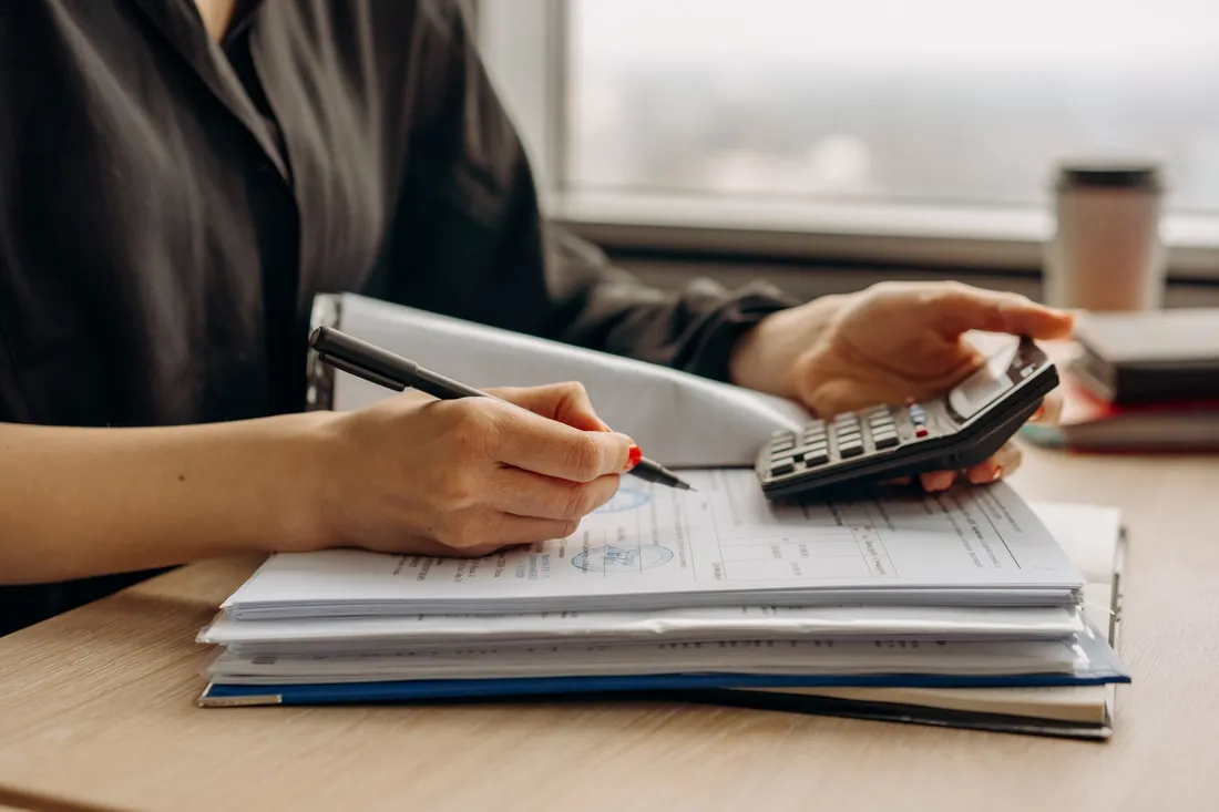 Personne utilisant une calculatrice, stylo à la main, au-dessus d'une pile de dossiers et de documents