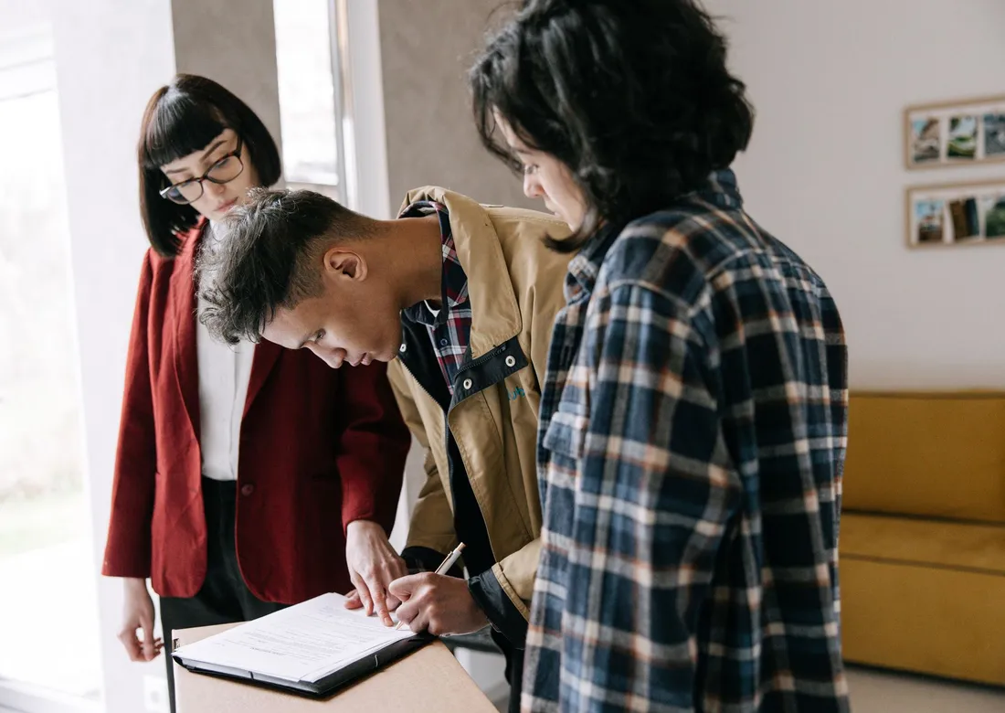 Un couple signe un contrat de vente immobilière sur un porte-documents, une agente immobilière à leurs côtés