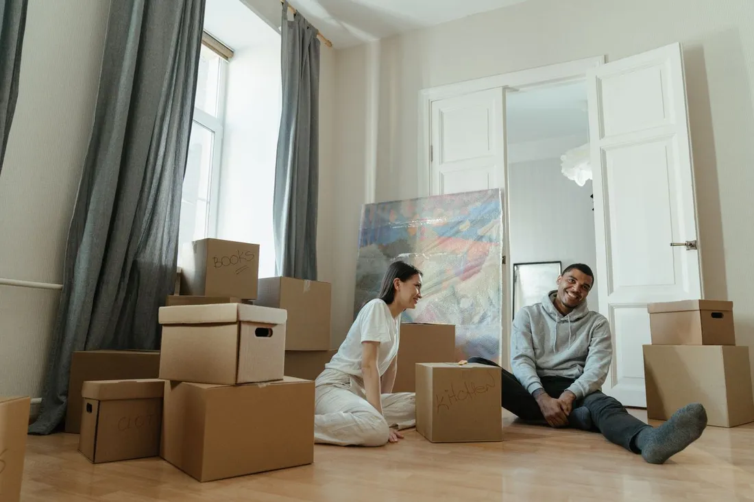 Jeune couple souriant assis parmi des cartons de déménagement étiquetés dans le salon lumineux de leur nouveau logement