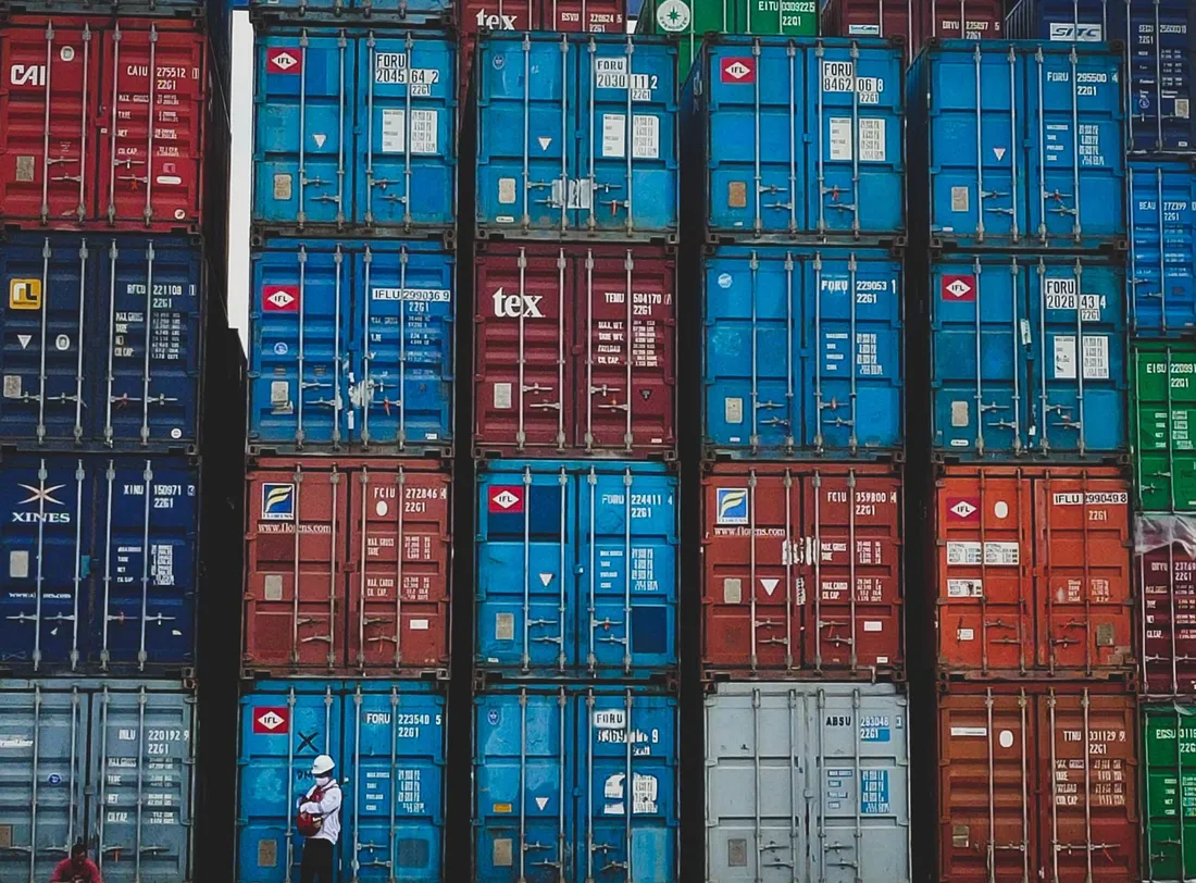 Conteneurs maritimes empilés sur plusieurs hauteurs dans un terminal portuaire, un docker en casque blanc au pied de la pile