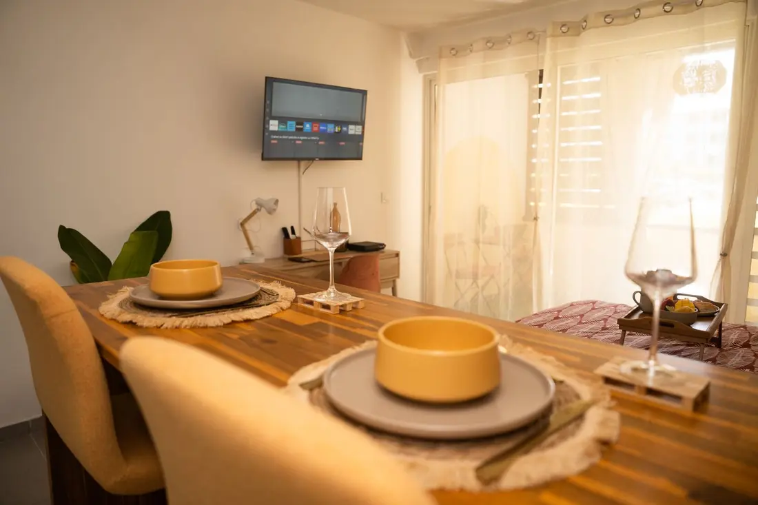 Table dressée du Cocon Chic : bols jaunes sur sets en fibres, verres à pied, téléviseur mural et lit en arrière-plan