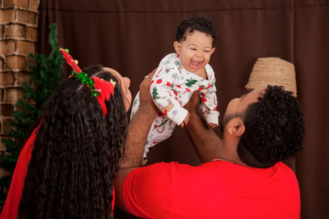 Famille caribeenne joyeuse celebrant Noel, soulevant un bebe en pyjama de fete