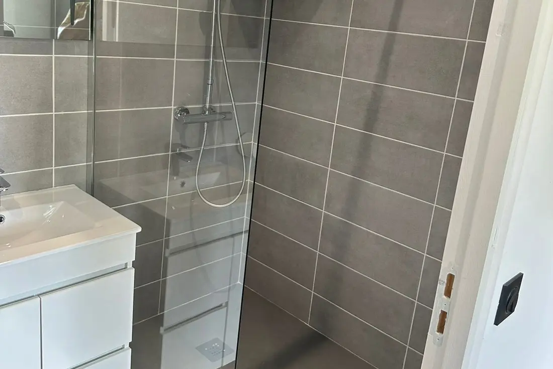 Salle d’eau d’un logement de la Résidence Francis : douche à l’italienne carrelée de gris et vasque blanche sur meuble laqué