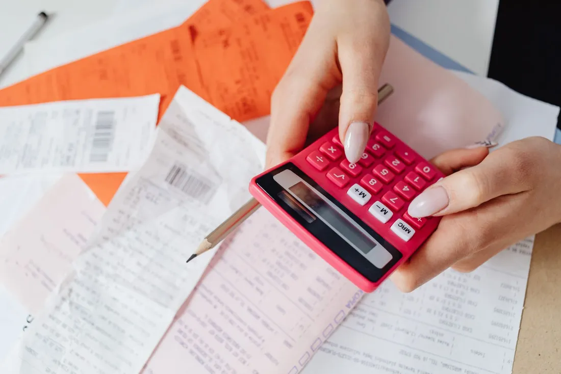 Mains vérifiant à la calculatrice une pile de tickets de caisse et de factures papier posés sur un bureau, comme lors de la préparation d'une note de frais