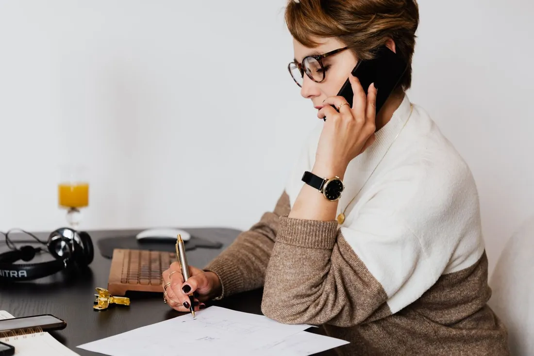 Femme à lunettes au téléphone à son bureau, un stylo à la main, annotant un document posé devant elle