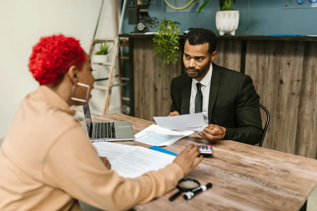 Rendez-vous de conseil dans un bureau : une femme de dos consulte un document et une calculatrice pendant qu'un homme en costume, assis face à elle, lui présente une autre feuille, un ordinateur portable ouvert entre eux