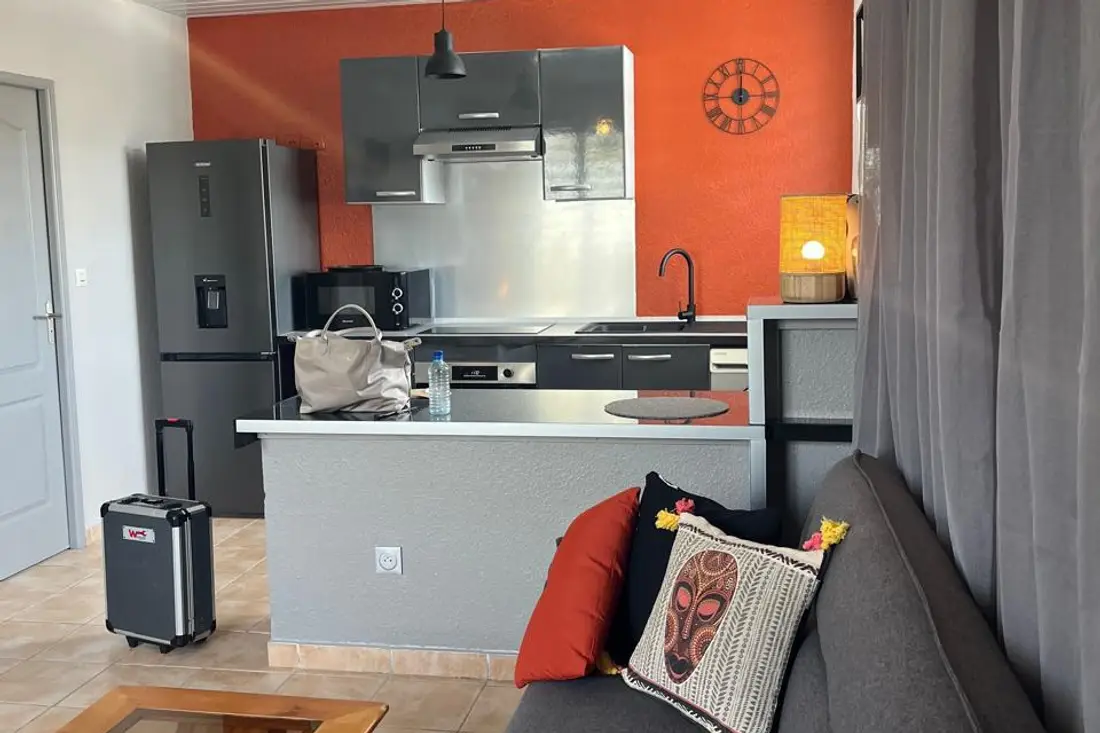 Séjour d’un appartement Cayenne Chic : valise à roulettes posée près du comptoir de la cuisine ouverte au mur orange