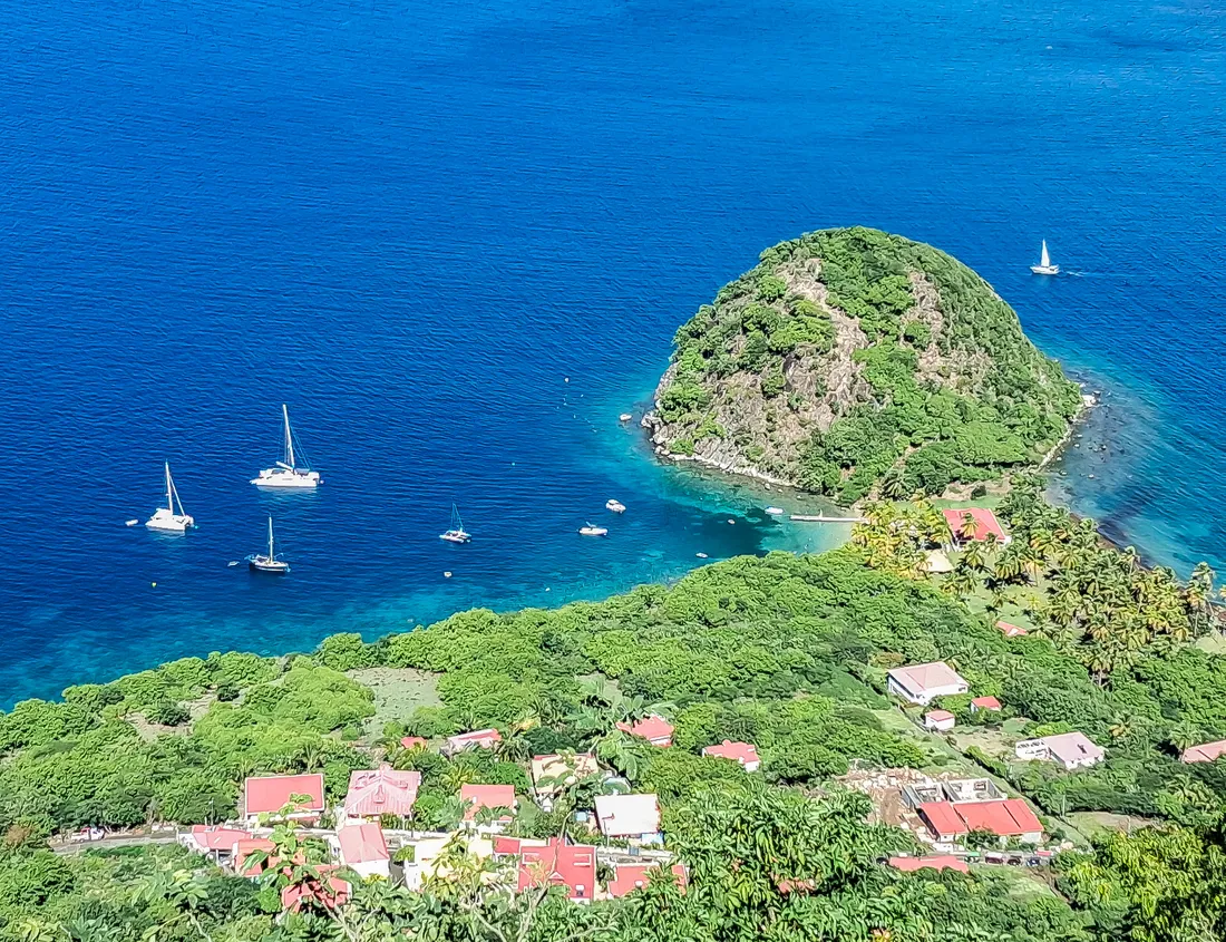 Le Pain de Sucre des Saintes, piton rocheux verdoyant cerné d'eau bleue, avec voiliers au mouillage et la crique abritée à ses pieds