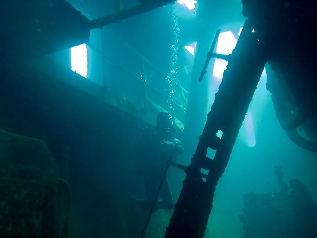 Plongeur à l'intérieur de l'épave du Franjack en Guadeloupe, gravissant une structure métallique avec la lumière filtrant par les ouvertures de la coque
