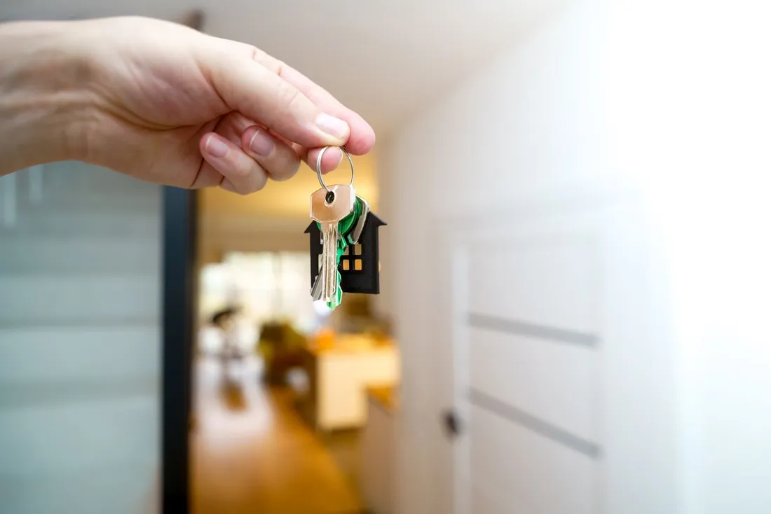 Main tendant un trousseau de clés avec porte-clés en forme de maison, devant l'entrée lumineuse d'un appartement