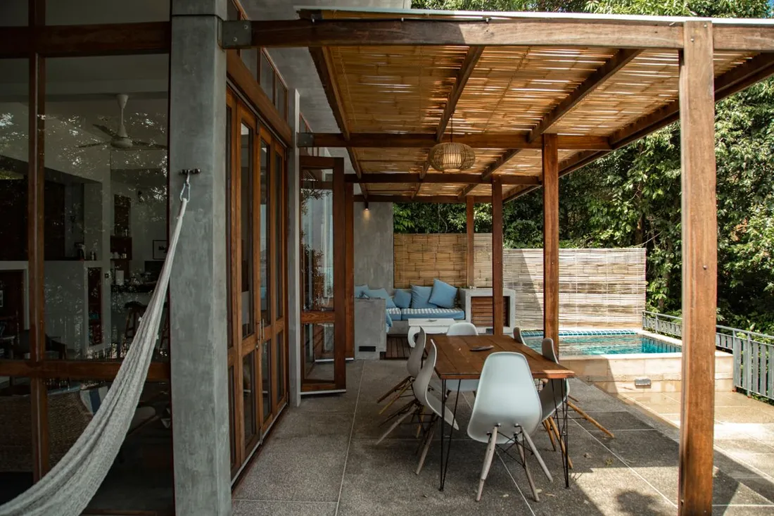 Terrasse couverte d'une location saisonnière tropicale avec hamac, table à manger et piscine entourée de verdure