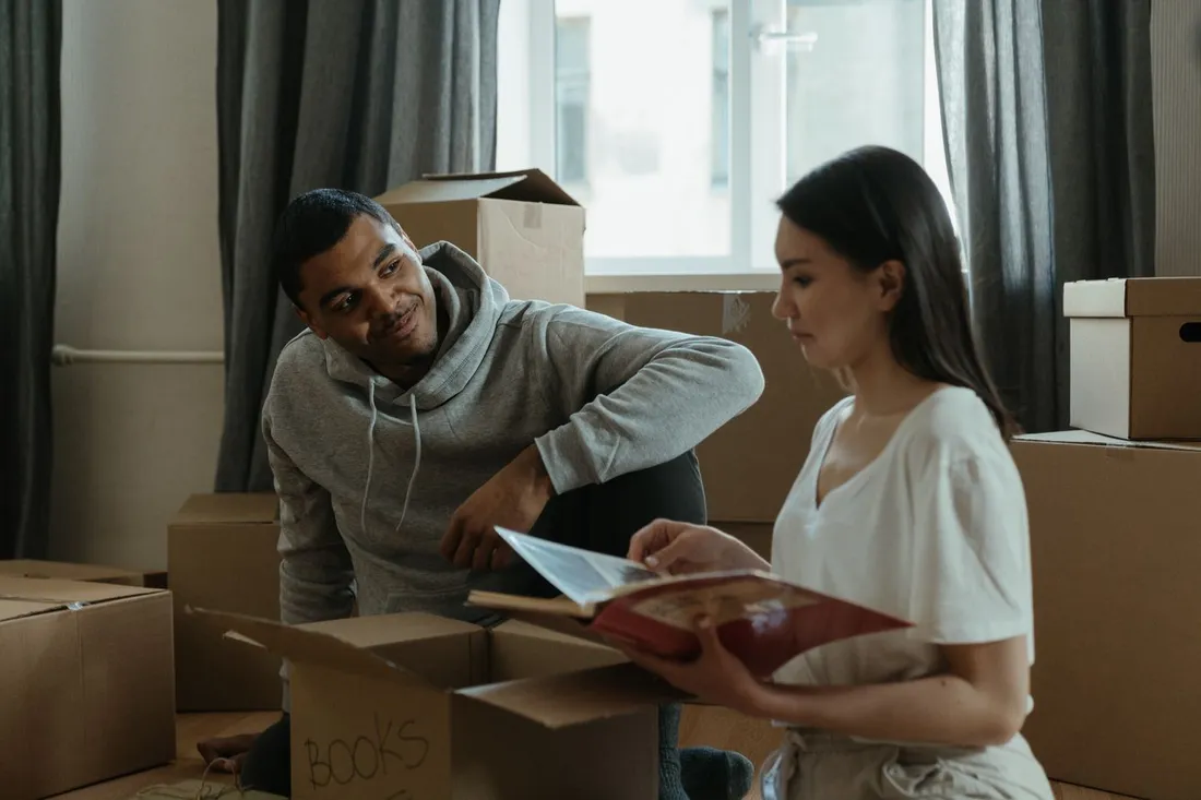 Un couple installe ses cartons de déménagement dans un appartement lumineux, feuilletant un livre au milieu des boîtes fraîchement ouvertes
