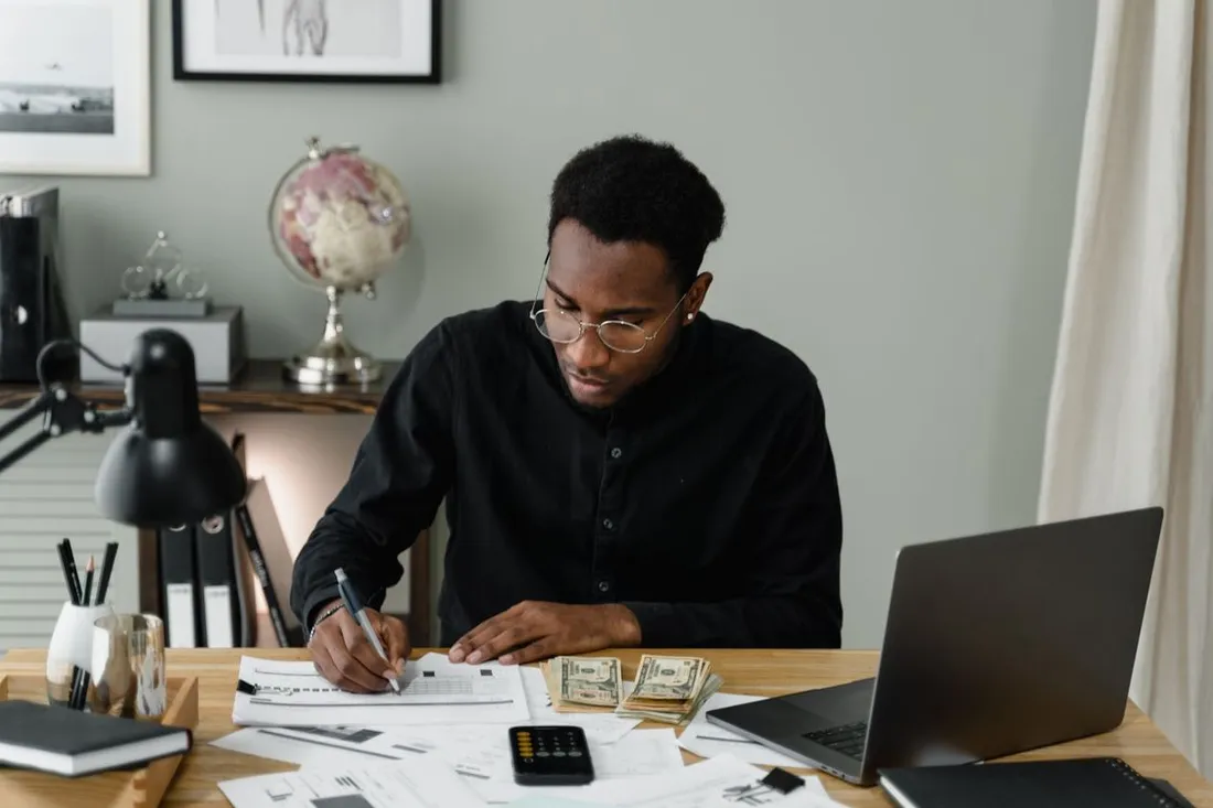Homme assis à un bureau, stylo à la main, annotant des relevés papier à côté d'un ordinateur portable, d'une calculatrice et de billets de banque