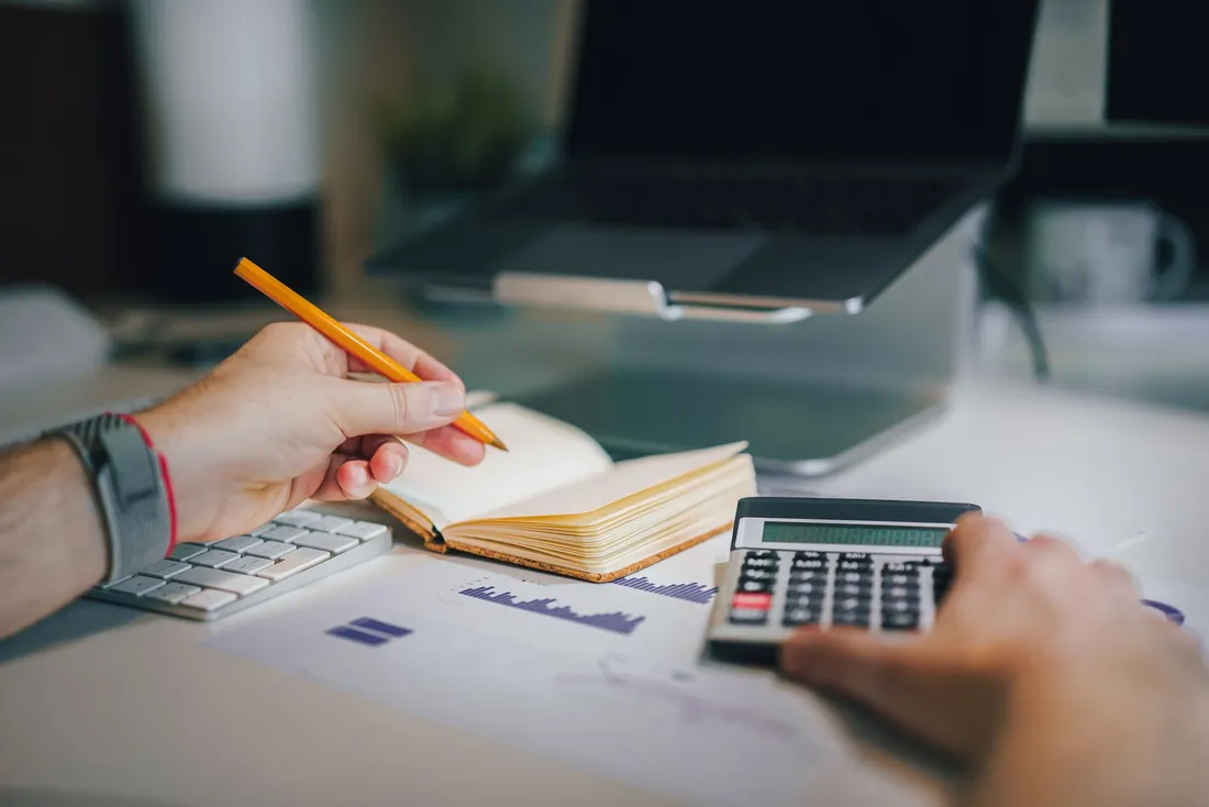 Deux mains sur un bureau : l'une tient un crayon au-dessus d'un carnet ouvert, l'autre tape sur une calculatrice posée à côté d'un graphique imprimé, devant un ordinateur portable
