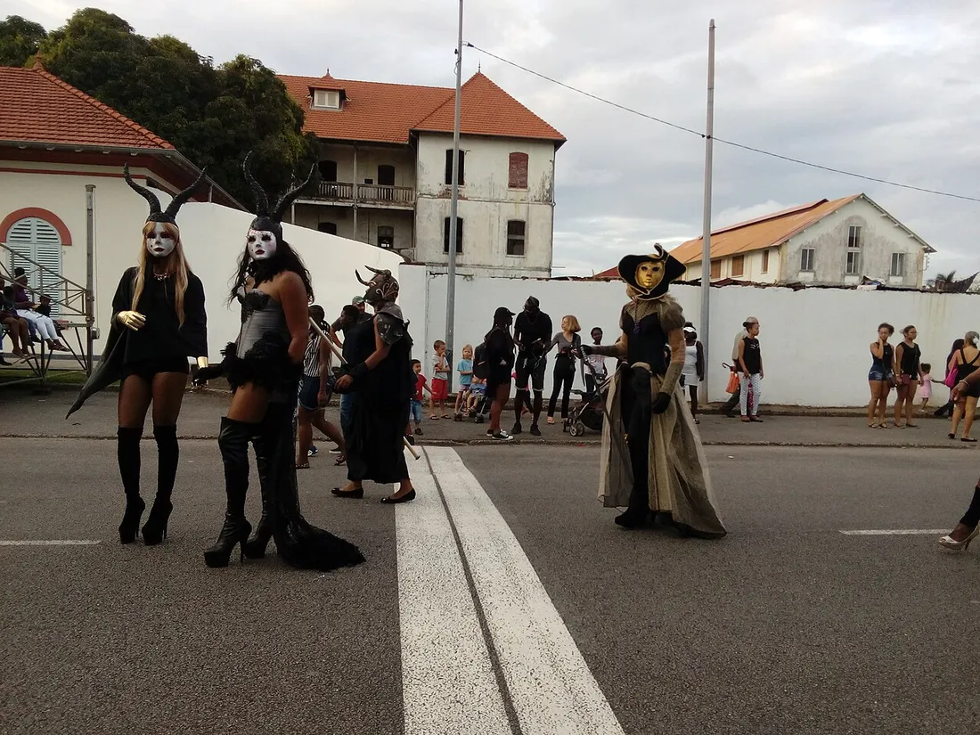 Défilé du carnaval dans une rue de Cayenne : trois personnes costumées et masquées avancent sur la chaussée devant des spectateurs et des maisons créoles à toiture rouge