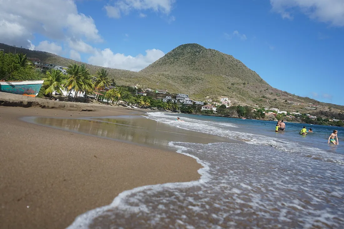 Plage de sable de l'Anse à l'Âne aux Trois-Îlets en Martinique, avec baigneurs, cocotiers et morne verdoyant en arrière-plan sous un ciel bleu