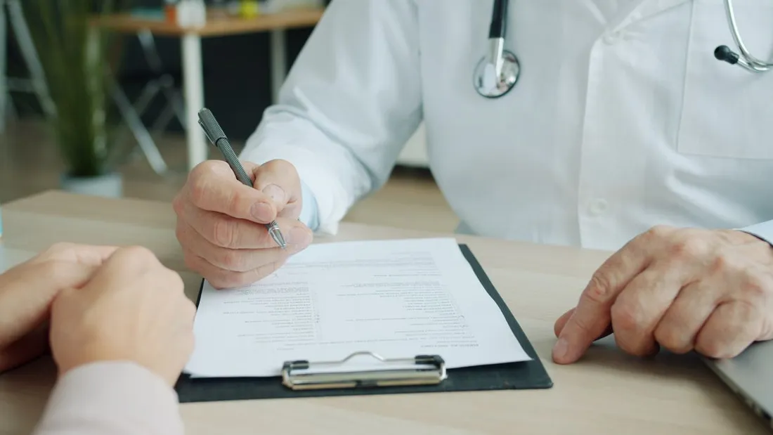 Médecin en blouse blanche avec stéthoscope remplissant au stylo un document fixé sur un porte-bloc, face à un patient assis de l'autre côté du bureau