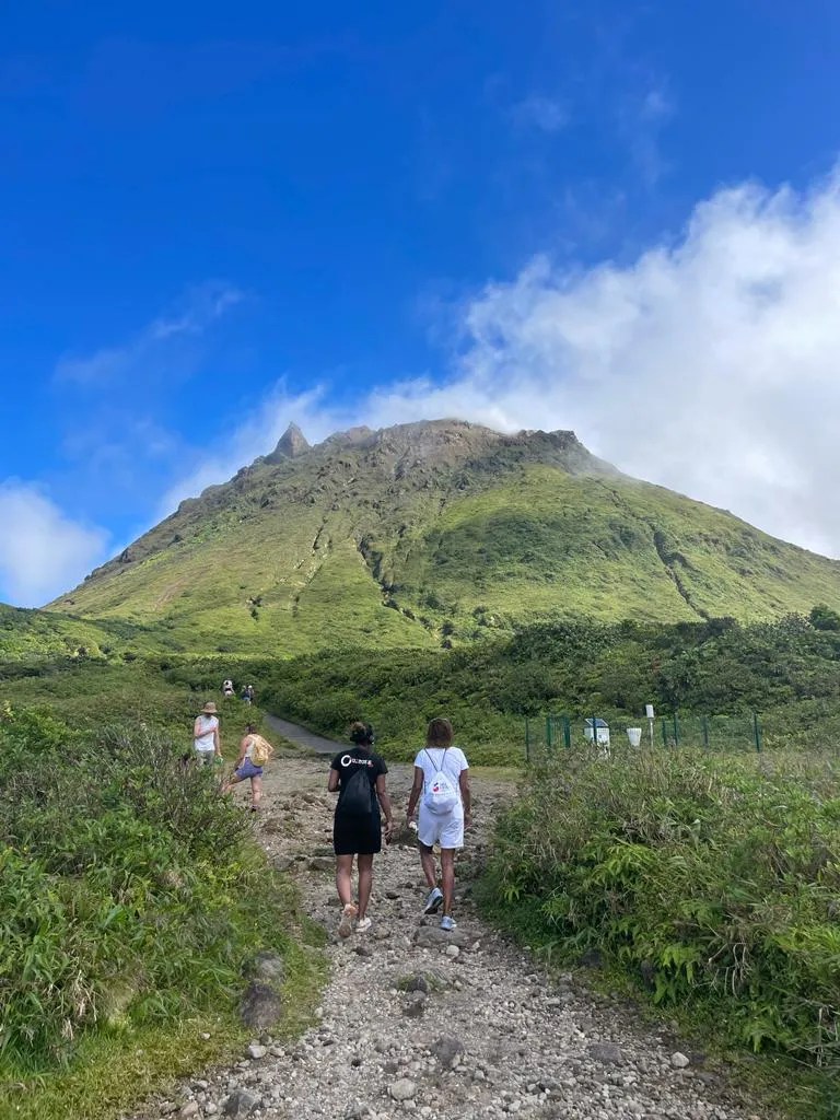Randonneurs gravissant le sentier rocailleux vers le sommet de la Soufrière en Guadeloupe, sous un ciel bleu et la brume accrochée au volcan