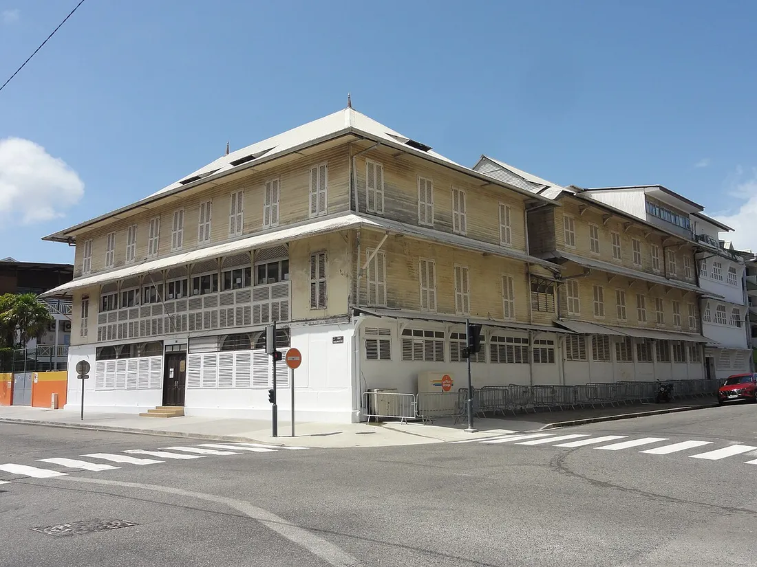 Le bâtiment en bois à deux étages de l'Externat Saint-Joseph de Cluny, à l'angle de deux rues du centre de Cayenne : volets persiennes, larges auvents et toiture de tôle claire sous un ciel bleu