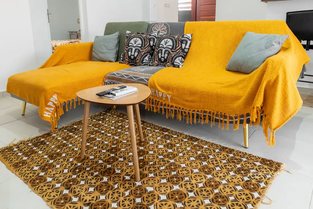 Séjour du Nid de Thalys : canapé couvert de plaids jaune moutarde, coussins à motifs de masques, table basse en bois