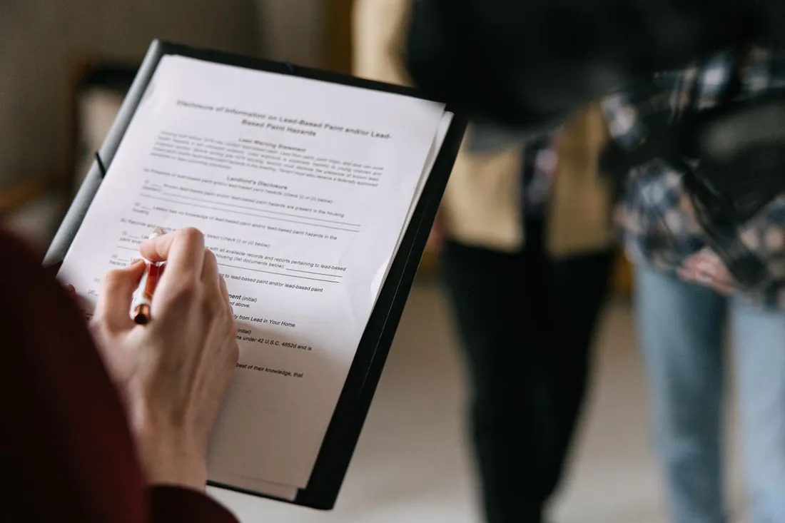 Une personne remplit au stylo un formulaire de location fixé sur un porte-documents, pendant que deux visiteurs discutent en arrière-plan dans un appartement