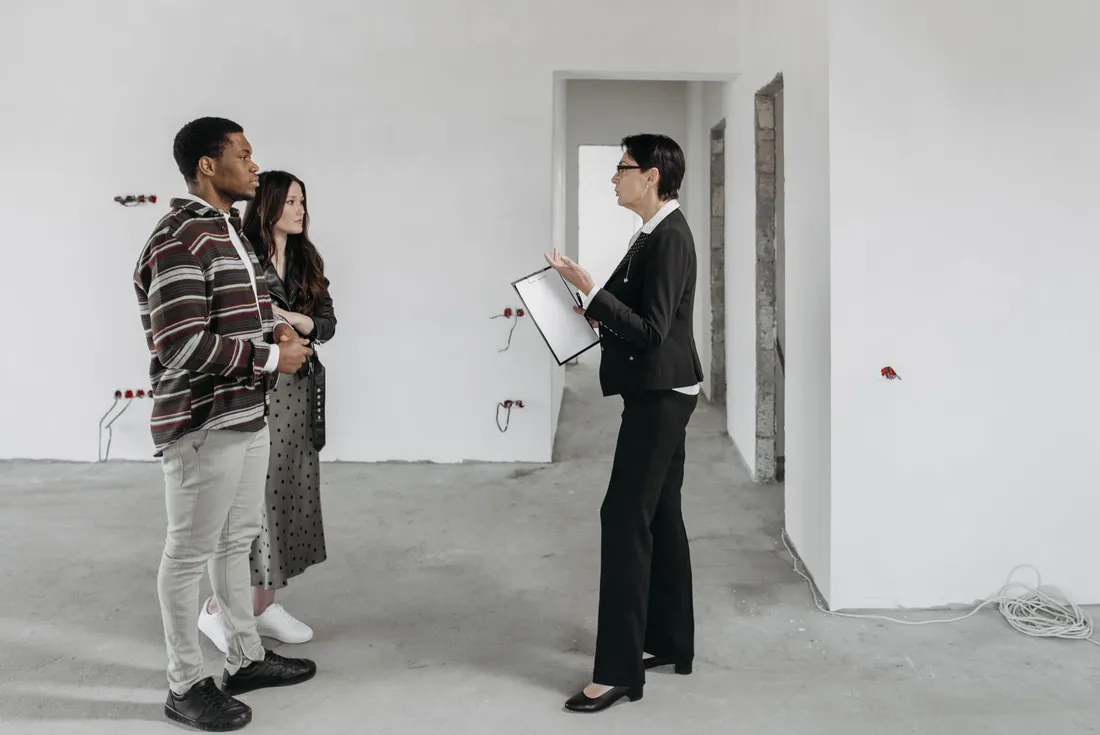 Un couple visite un logement neuf non fini, murs blancs bruts et gaines électriques apparentes, face à une conseillère en tailleur noir tenant un porte-documents