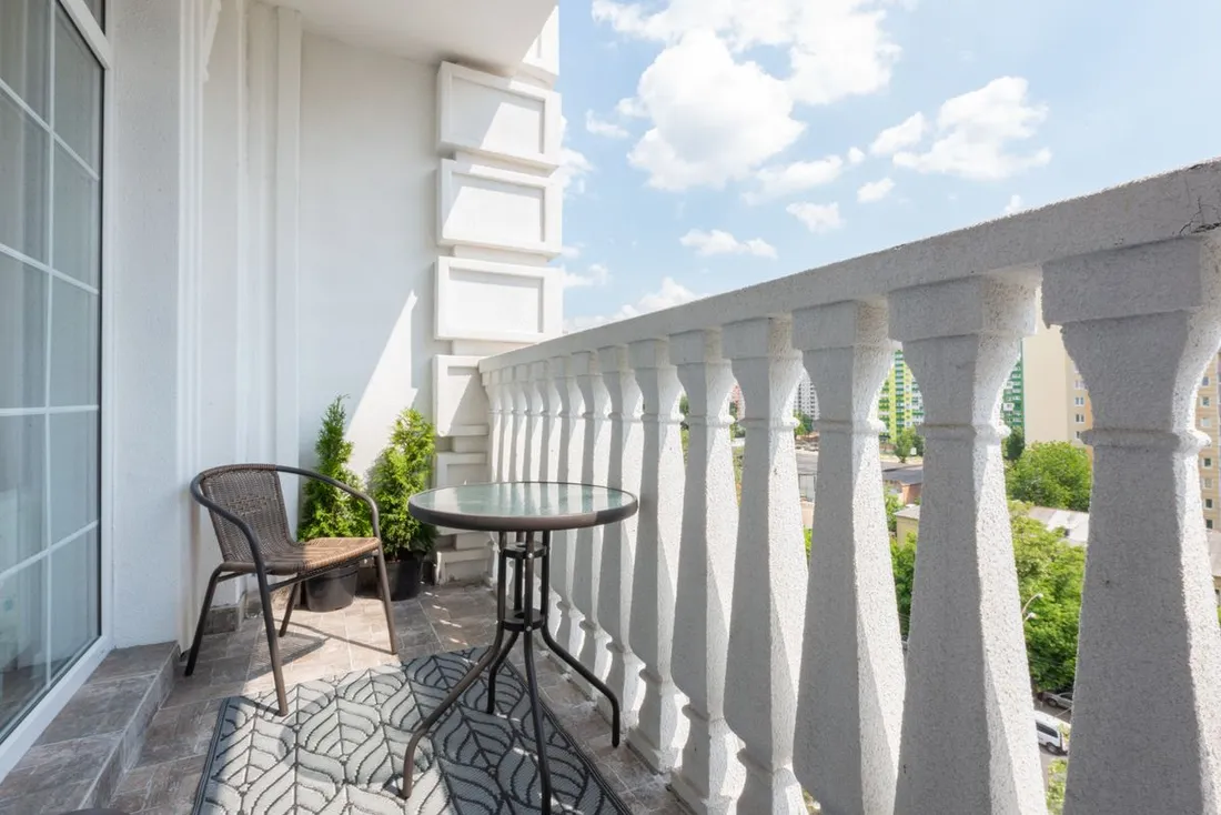 Balcon d'appartement avec une chaise en résine tressée, une table ronde en verre et un tapis, derrière une balustrade blanche à colonnettes ouvrant sur les arbres et les immeubles voisins
