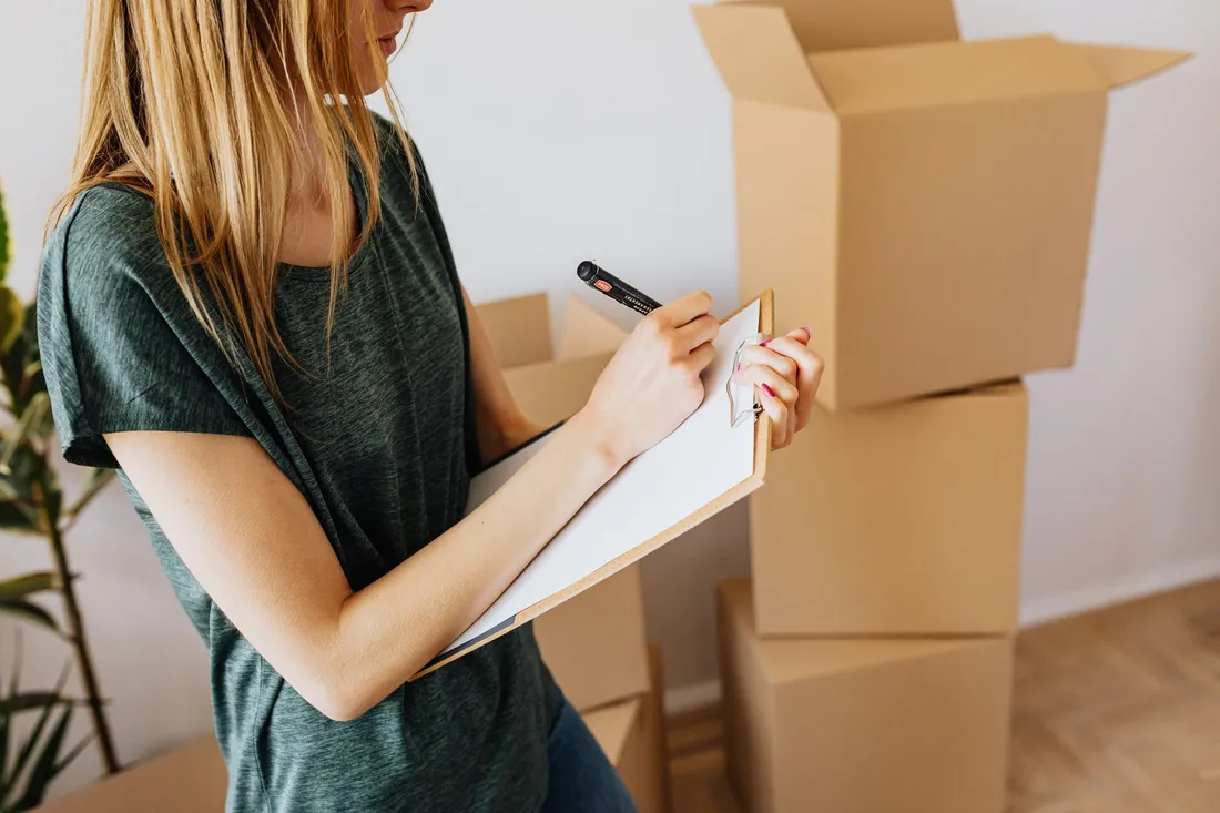 Une femme en tee-shirt, debout dans un logement aux murs blancs, note quelque chose sur une planchette à pince ; derrière elle, des cartons de déménagement empilés et un carton encore ouvert