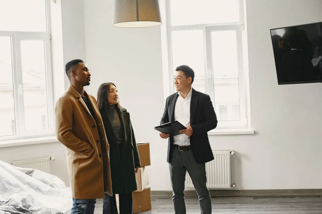 Un agent immobilier, planchette à la main, présente un appartement vide à un couple debout au milieu de cartons de déménagement