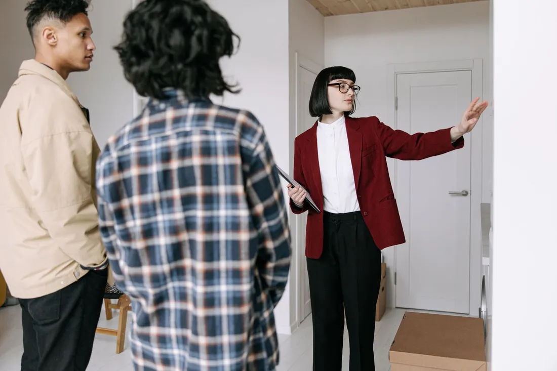 Une agente en veste rouge, tablette à la main, désigne une pièce à un couple de visiteurs dans un appartement en cours d'emménagement